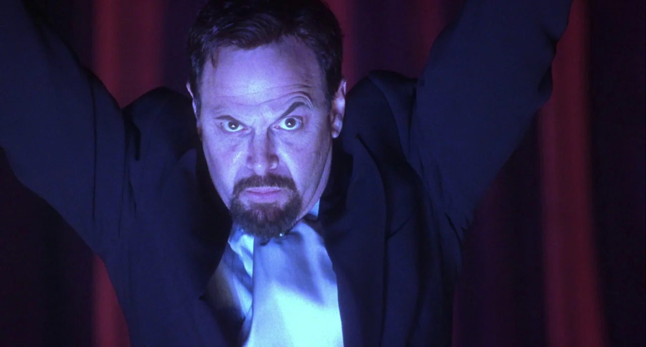 Mulholland Drive (2001) movie still: closeup — A man stands in front of dark red velvet curtains on a dimly lit stage. The man reaches his arms…