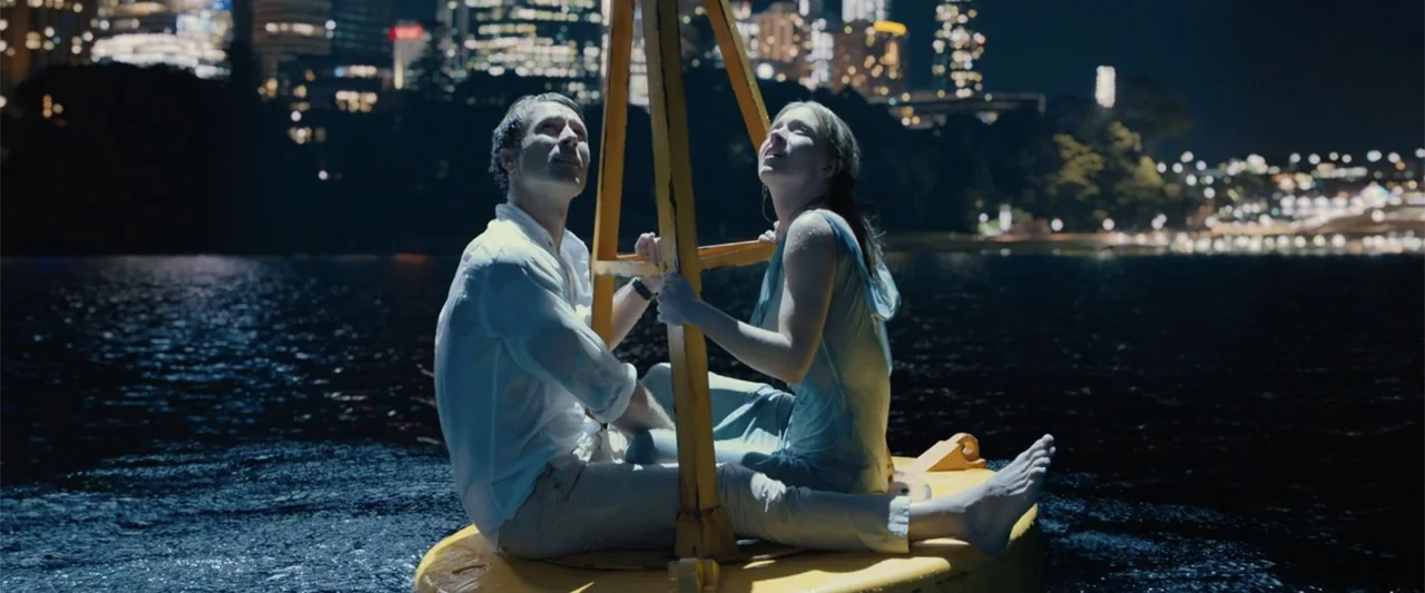 Anyone But You (2023) movie still: medium — A bright yellow navigational buoy floats in dark water at night near a city. A man and a woman sit atop…