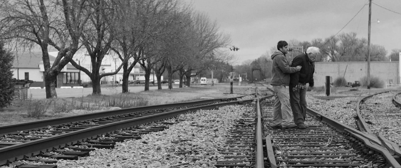 Nebraska (2013) movie still: wide — A set of railroad tracks stretches through a rural landscape with bare autumn trees. A younger man assists an…