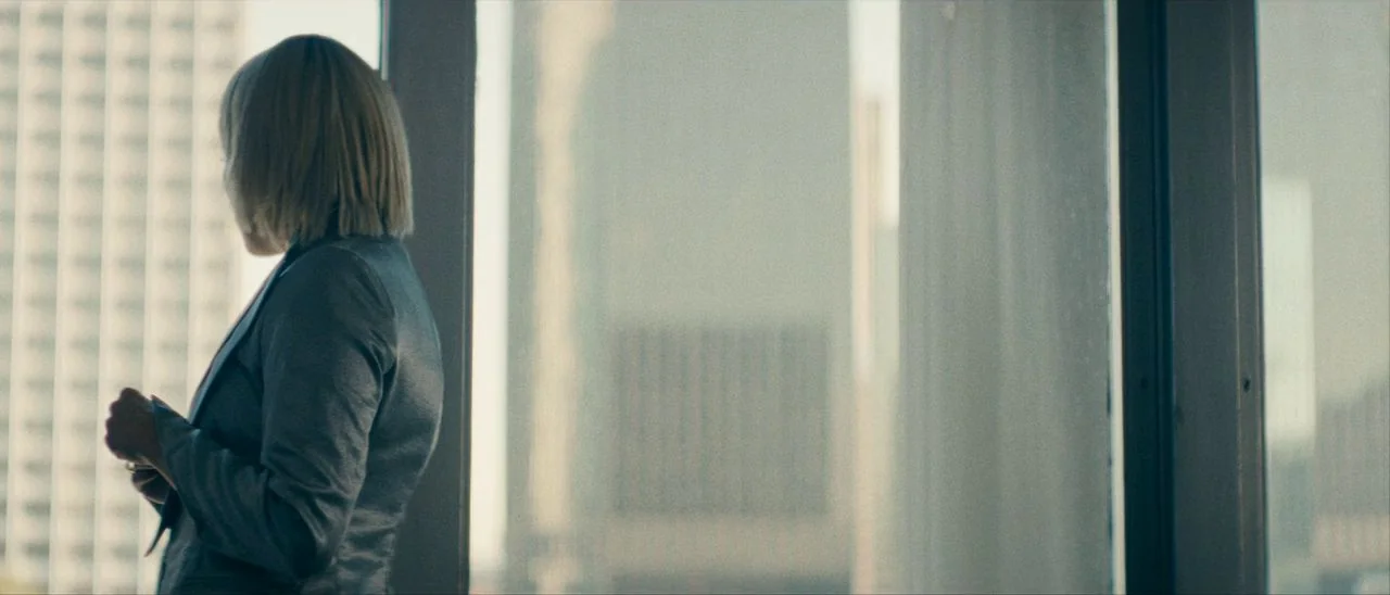 Archenemy (2020) movie still: medium — A high-rise room with expansive windows overlooks a dense metropolitan area of tall buildings. A blonde woman…