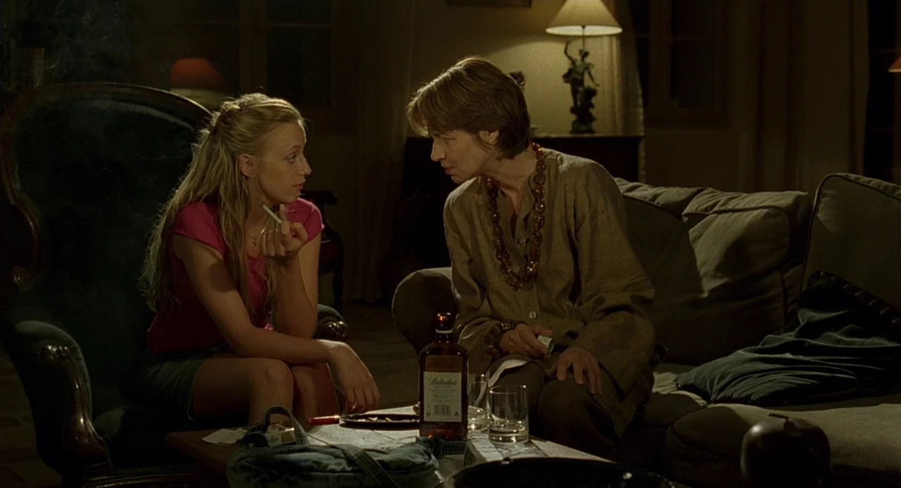 Swimming Pool (2003) movie still: medium — A dark, dimly lit domestic space filled with soft shadows and furniture. Two women converse while sitting…