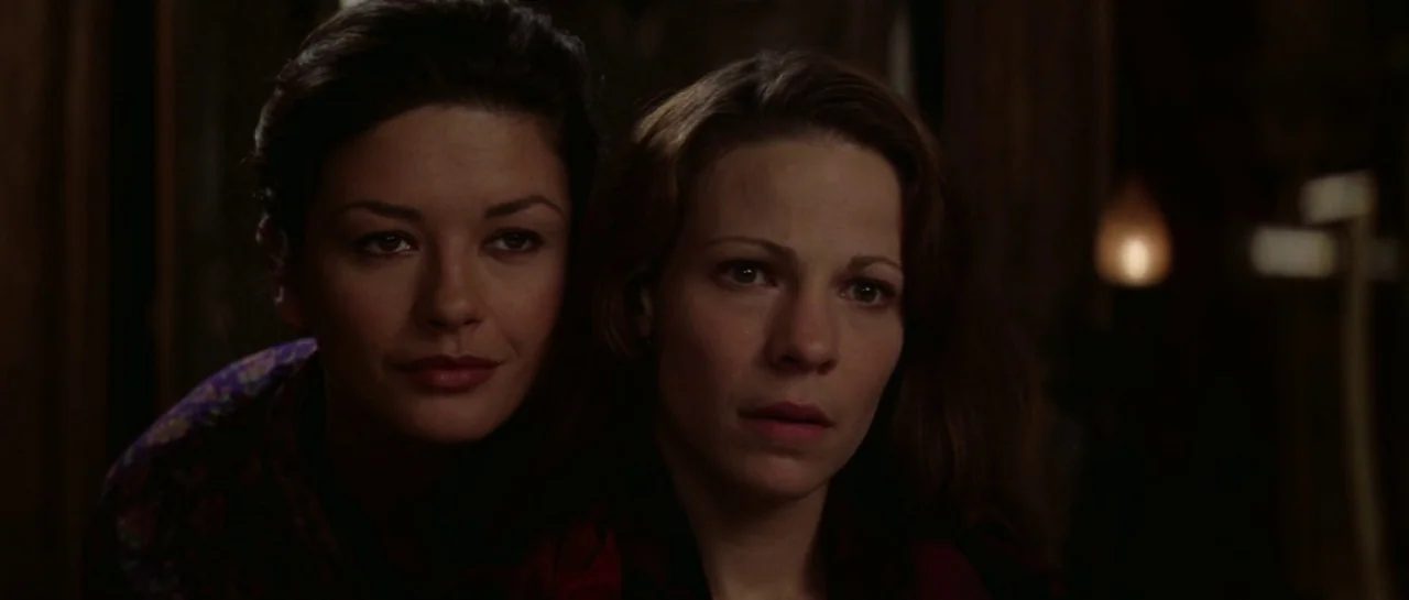 The Haunting (1999) movie still: closeup — Two women stand together in a dark room illuminated by a distant flame light. The women gaze forward…