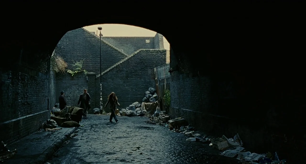 Children Of Men (2006) movie still: wide — A dark, arched brick underpass contains scattered trash and debris on a wet ground surface. A woman walks…