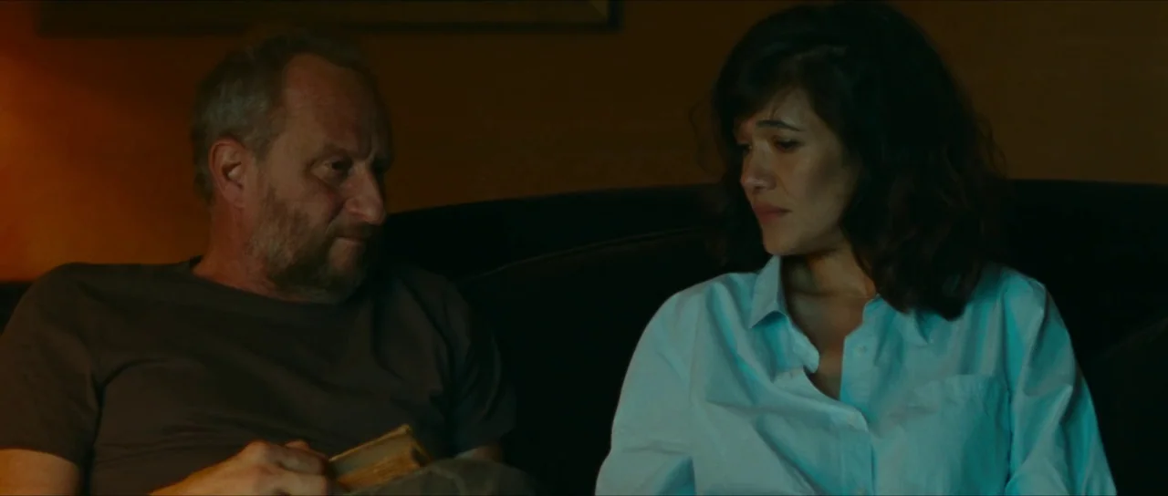 Inexorable (2021) movie still: medium — A dimly lit room with a man and woman sitting on a dark couch. The man holds an open book while the woman…