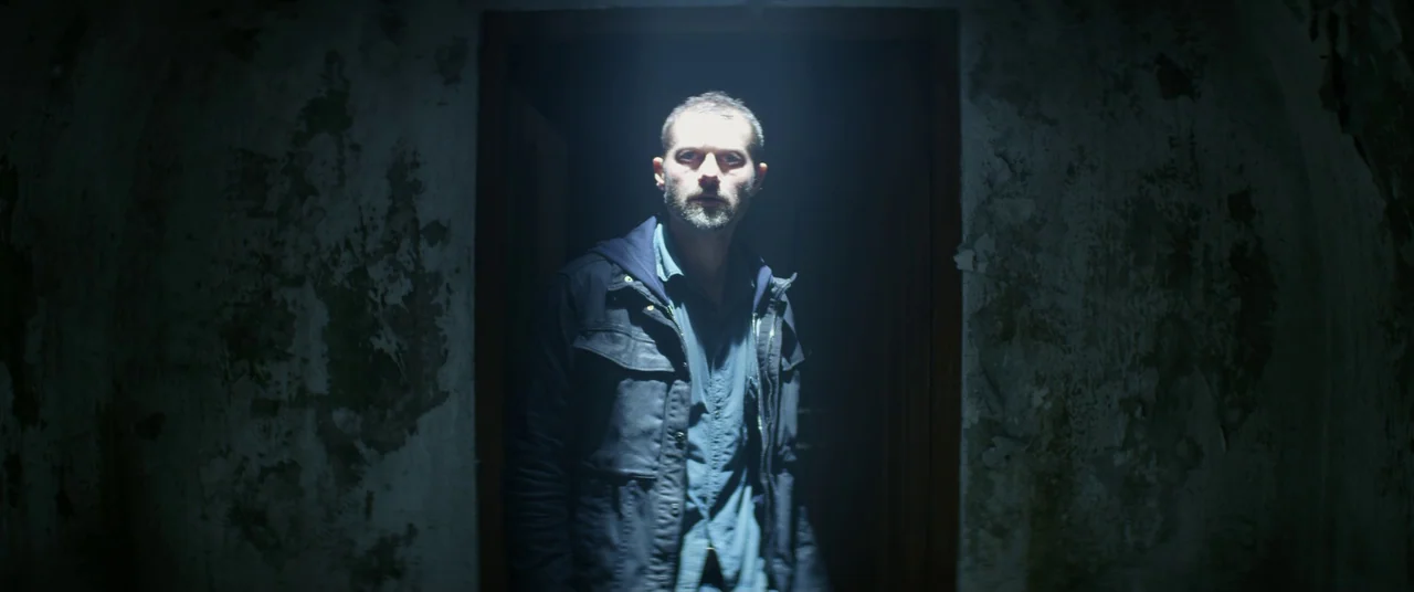 The Empty Man (2020) movie still: medium — A man stands in a dark, narrow doorway flanked by crumbling, textured plaster walls. He is illuminated by…