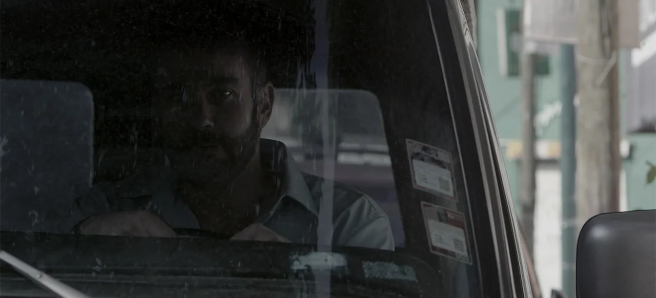 About the Deaths (2016) movie still: closeup — A view through a car windshield shows a man sitting in the driver's seat. Stickers are visible on the…