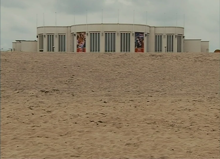 The Beaches of Agnès (2008) movie still: establishing — A wide sandy beach stretches toward a large modern building with a curved facade. The…
