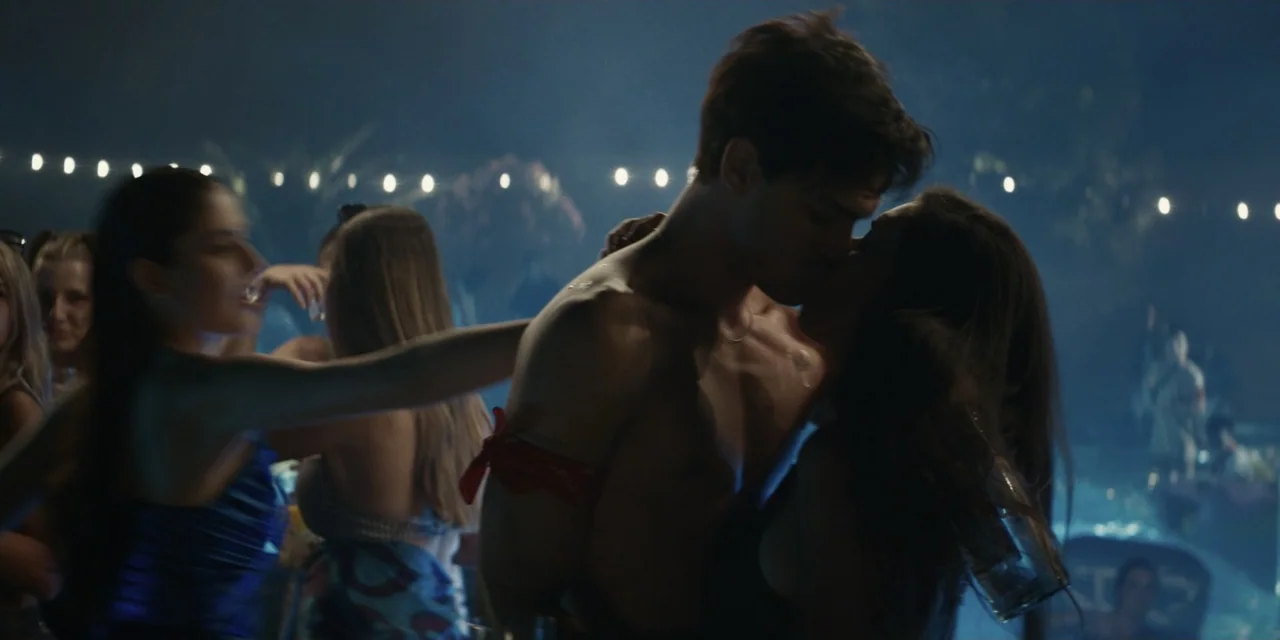 My Fault (2023) movie still: medium — An outdoor night party setting features string lights hanging above a crowd of people. A man and a woman stand…