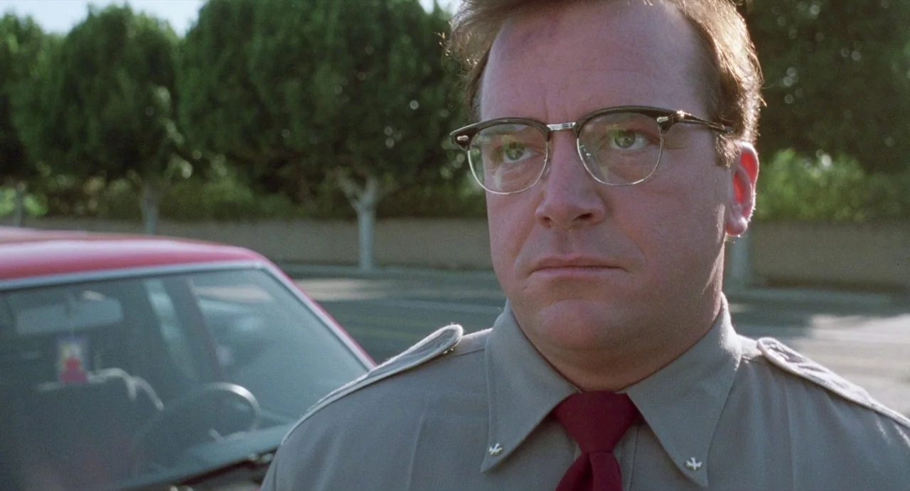 Touch (1997) movie still: medium — A sunny outdoor parking lot with trimmed green hedges in the background. A man wearing a uniform stands in the…