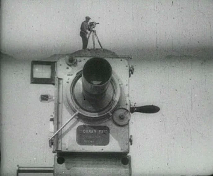 Man with a Movie Camera (1929) movie still: medium — A man stands atop a large Debrie Parvo cinema camera in an outdoor space. He looks through a…