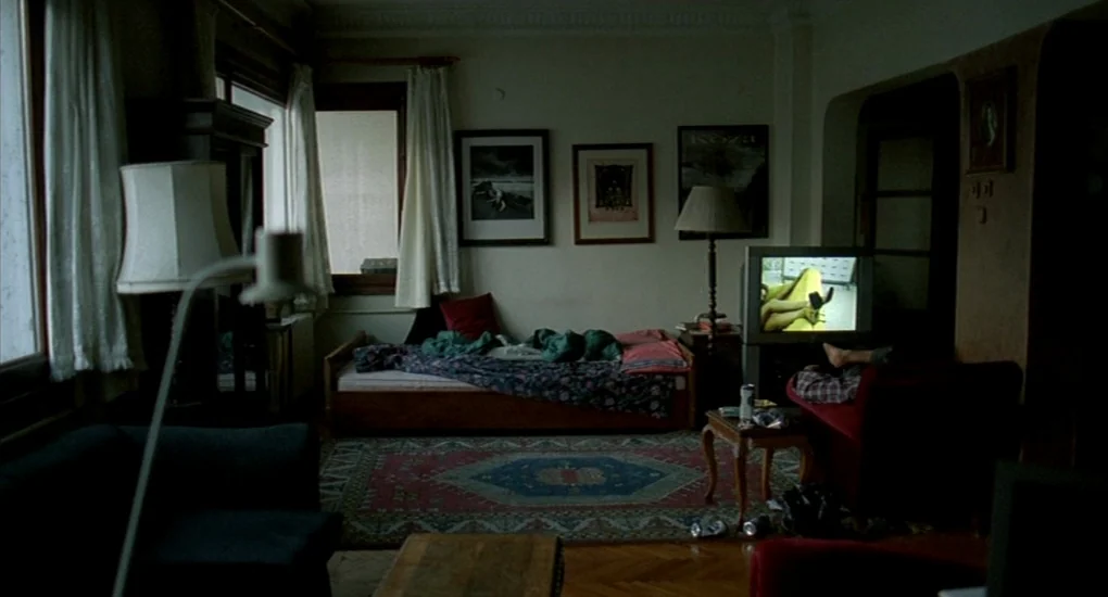 Uzak (2002) movie still: wide — A dimly lit room contains a patterned rug, a single bed, and a television. A pair of feet rests on the edge of a red…