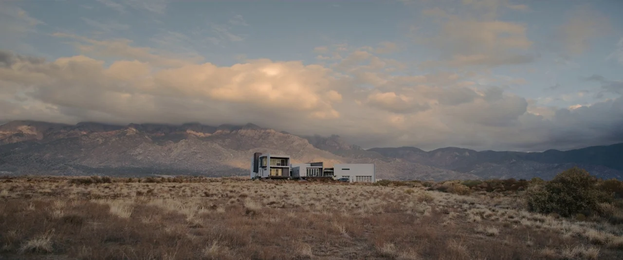 Intrusion (2021) movie still: establishing — A modern multi-level house sits in the middle of a vast, dry desert field. Large jagged mountains under…