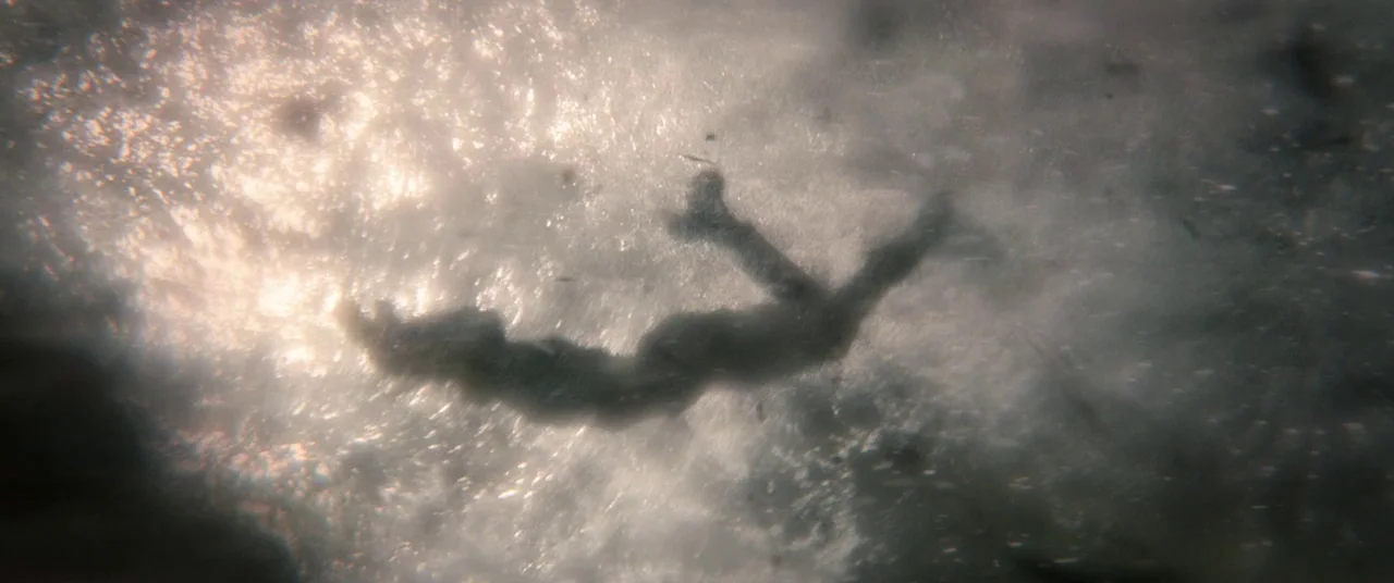 Tomb Raider (2018) movie still: wide — A dark silhouette of a person floats horizontally beneath a bright, rippled water surface. Sunlight filters…