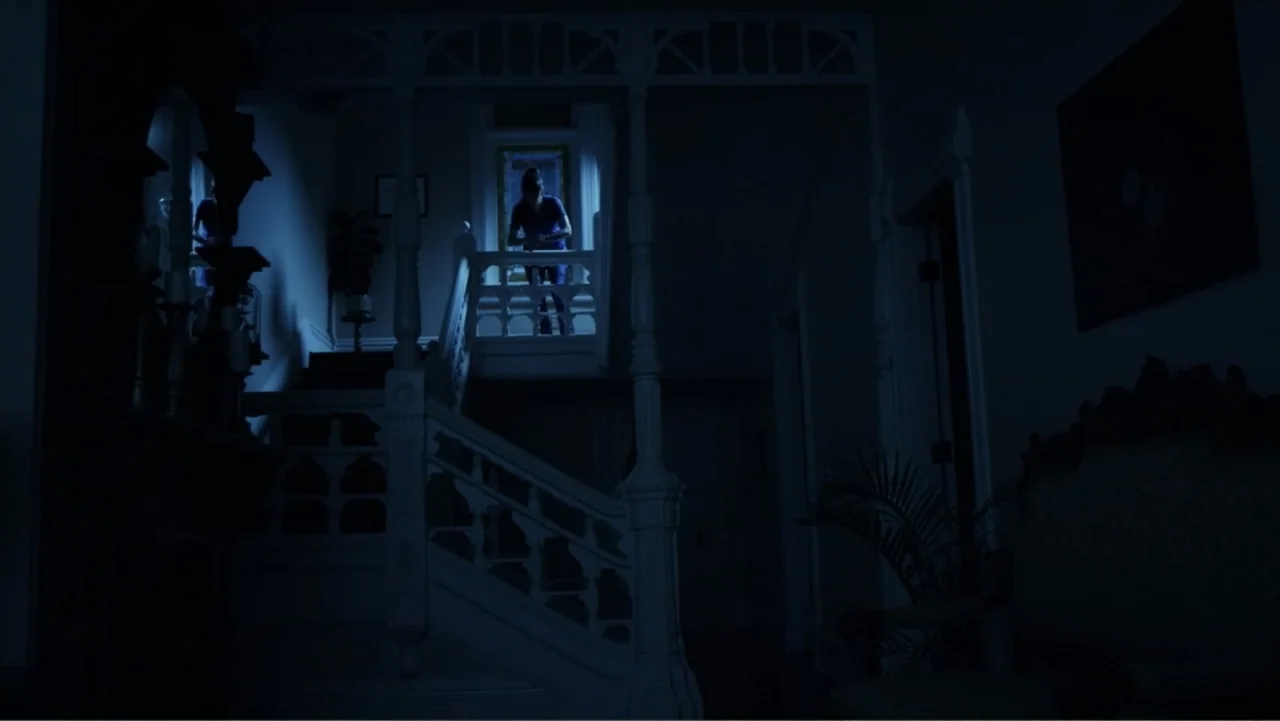 Storm Damage (2000) movie still: wide — A dark foyer features a traditional staircase and an ornate wooden banister. A single woman stands on the…