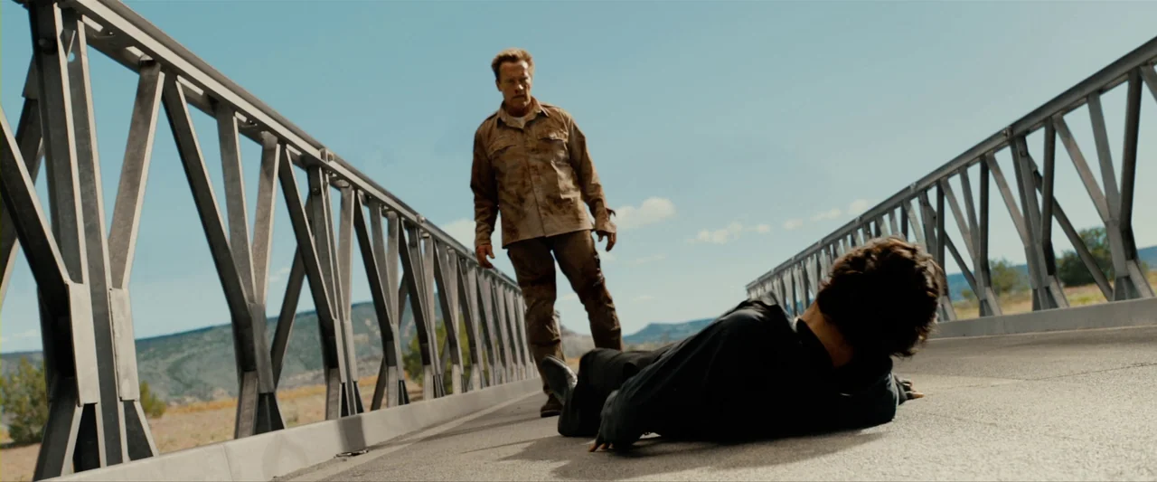 The Last Stand (2013) movie still: fullbody — A metal truss bridge stretches across a dry rural landscape under a clear sky. A man in stained tan…