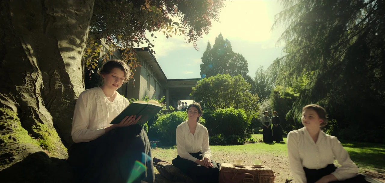 Peter Pan & Wendy (2023) movie still: wide — A sun-drenched lawn in front of an estate with trees and manicured shrubbery. Three young women sit on…