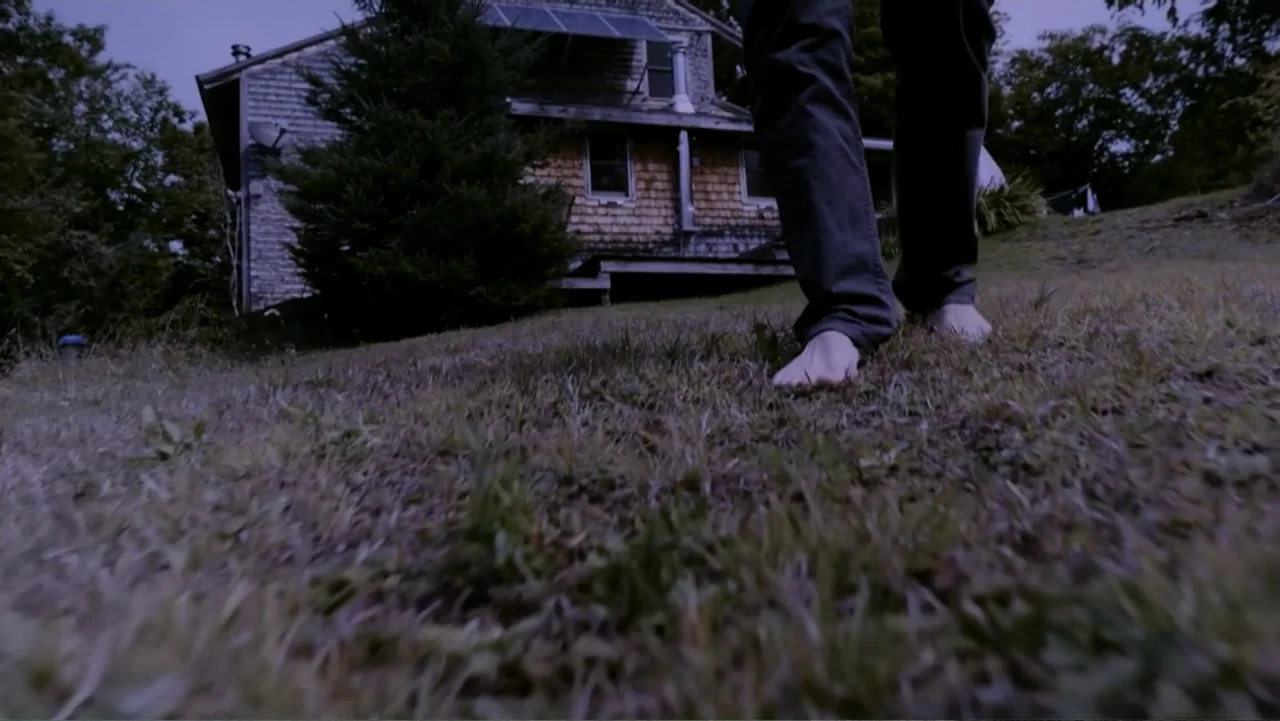 Beware of Angels (2016) movie still: medium — A dark house with shingled walls stands in the background behind an untrimmed lawn. A person walks…