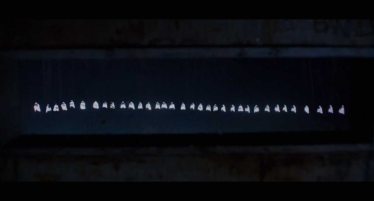 True History of the Kelly Gang (2019) movie still: wide — A dark, rectangular frame displays a long, uniform row of small, bright white figures. The…