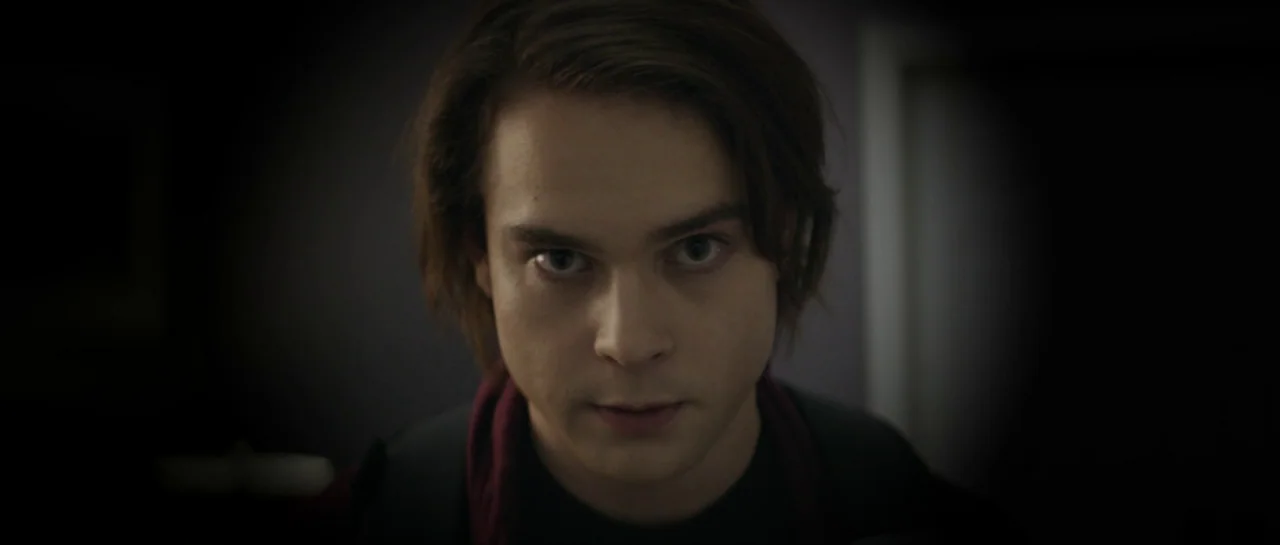 Suitable Flesh (2023) movie still: closeup — A dimly lit interior space with dark walls and heavy vignetting. A young man with brown hair stares…
