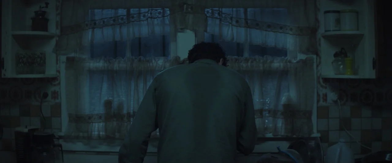 Wolf Man (2025) movie still: medium — A cluttered residential kitchen features patterned wallpaper and lace curtains over a small window. A lone man…