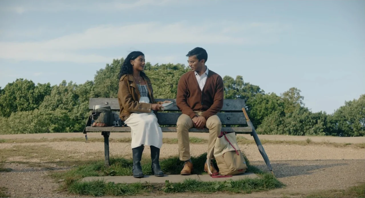 Picture This (2008) movie still: fullbody — An outdoor park setting features a wooden bench placed on a grassy gravel patch. A young woman and man…