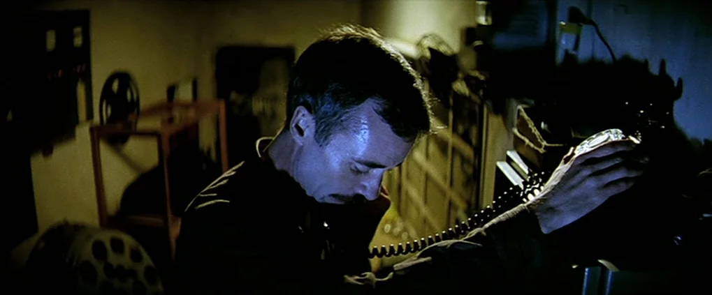 Anguish (1987) movie still: medium — A dimly lit room filled with film reels and technical equipment in shadows. A man with a mustache talks on a…