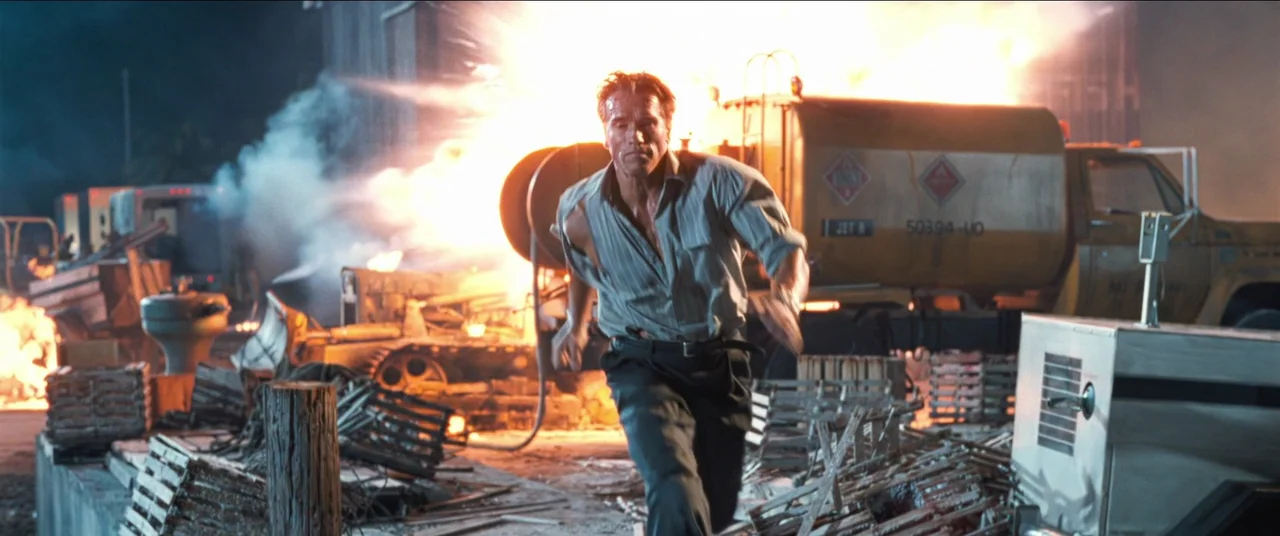 True Lies (1994) movie still: fullbody — An industrial outdoor space is filled with wooden crates and a large fuel tanker. A muscular man runs…