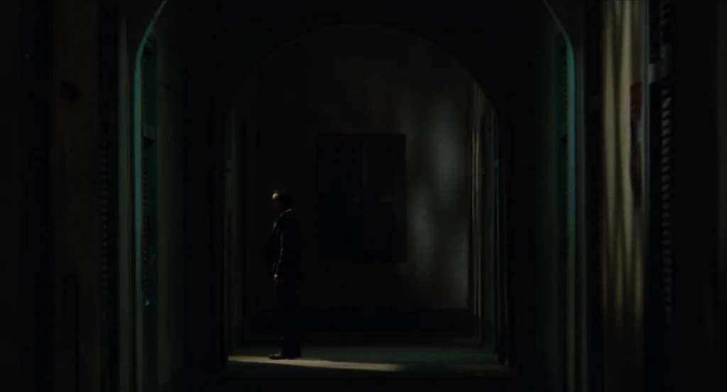 Almayer’s Folly (2011) movie still: wide — A dark, narrow interior hallway features arched doorways and shadowy walls. A lone man in a dark suit…