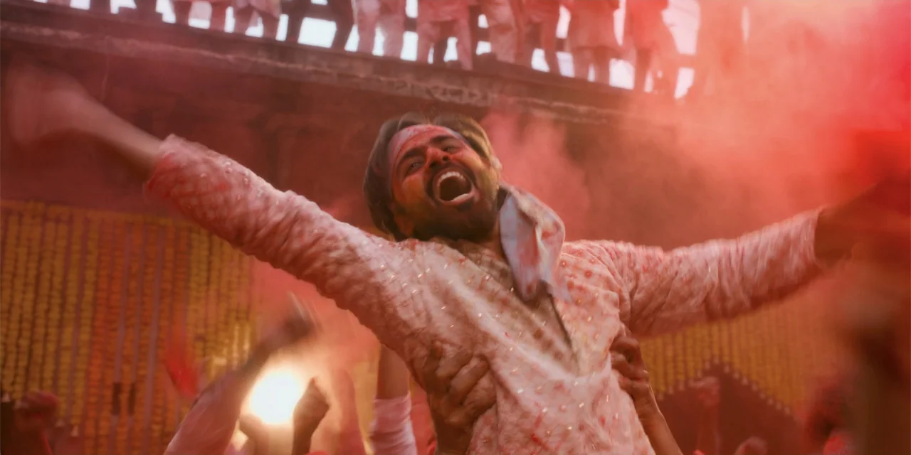 Dry Day (2023) movie still: medium — A man in a light pink kurta is lifted by a crowd during a Holi celebration. Bright red powder fills the air…