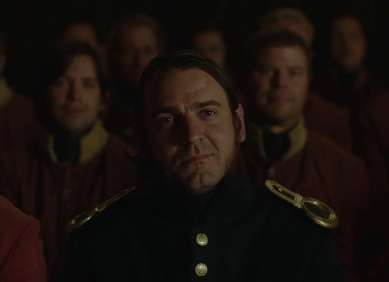 The Nightingale (2018) movie still: medium — A dark, wood-paneled room is filled with men in period military uniforms. A man in the foreground faces…