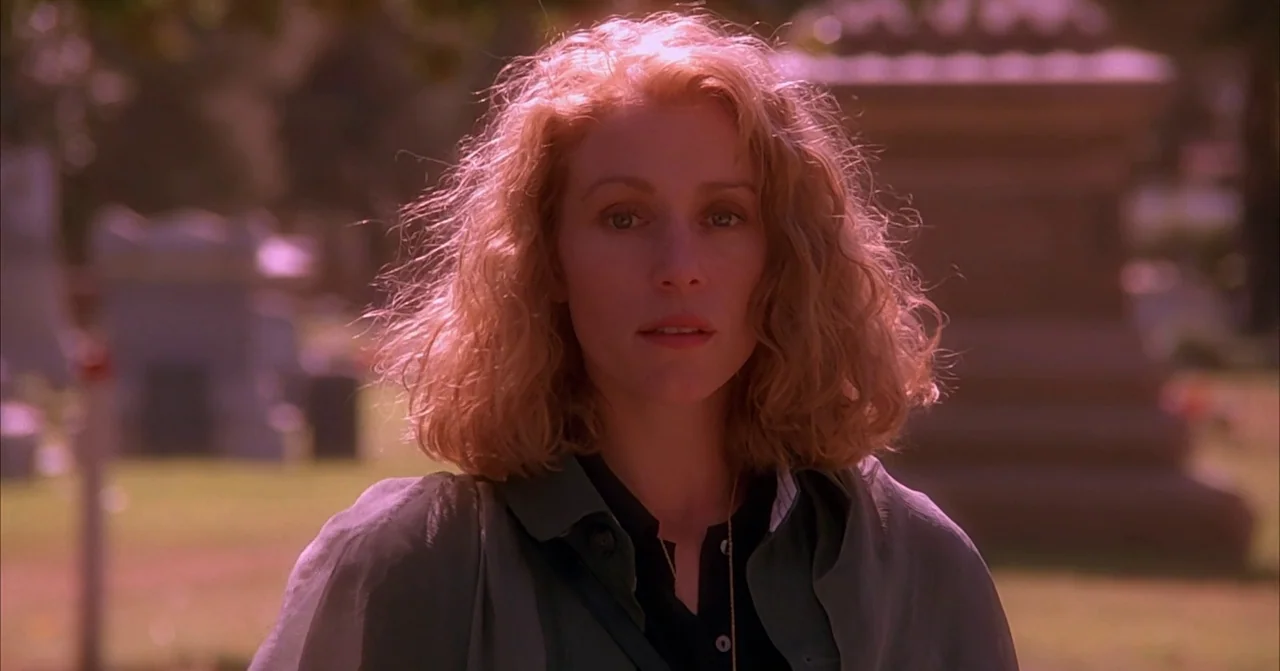 Darkman (1990) movie still: medium — A woman stands outdoors with gravestones blurred in the background. She faces the camera directly while…
