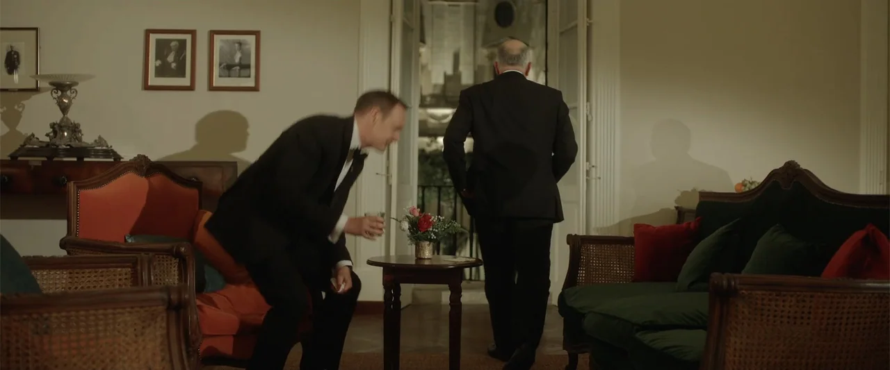 Hounds of War (2024) movie still: medium — A classic living room interior features upholstered armchairs, a small center table, and framed artwork.…