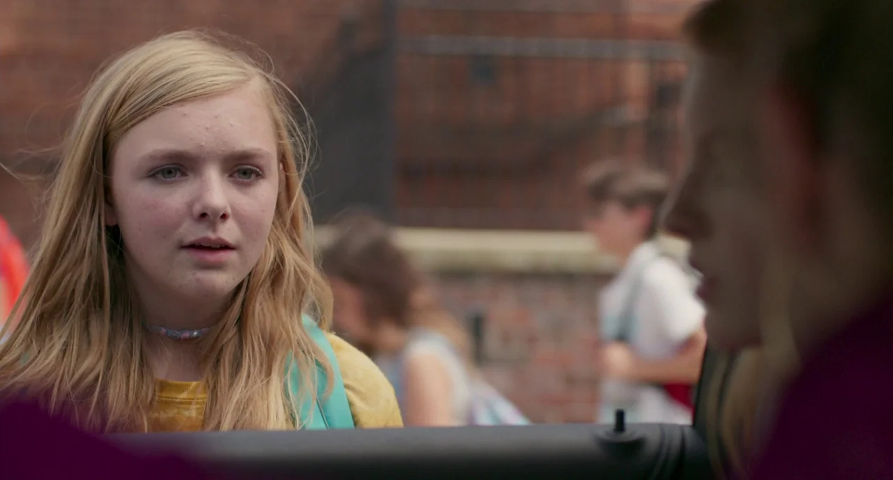 Eighth Grade (2018) movie still: medium — An outdoor area with a red brick wall and blurred figures in the background. A teenage girl talks to…