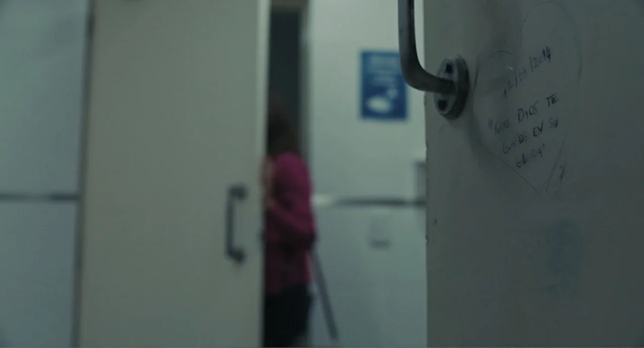 Belén (2025) movie still: medium — A public restroom stall door features handwritten text inside a drawn heart shape. A woman in a pink top stands…