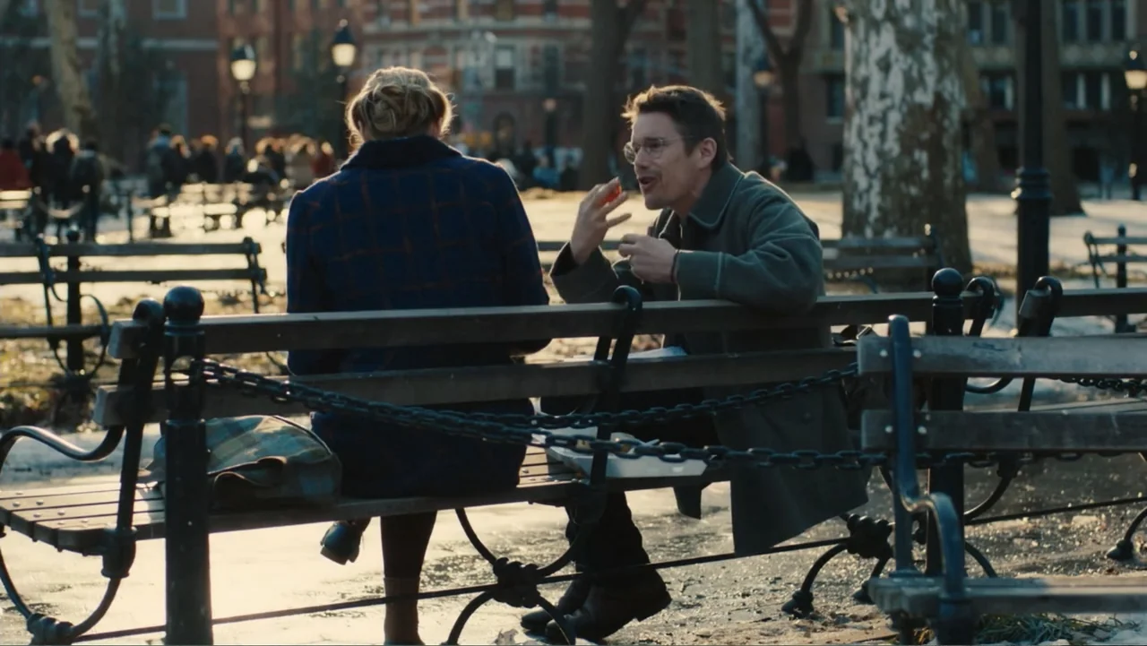 Maggie's Plan (2016) movie still: medium — A public park with bare trees and scattered snow on the ground at dusk. A couple sits together on a…