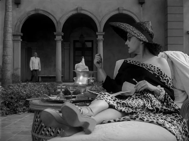 Sunset Boulevard (1950) movie still: medium — A stone patio with arched columns features a woman relaxing on a lounge chair. In the background a man…