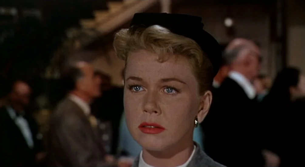 The Man Who Knew Too Much (1956) movie still: closeup — A crowded interior space with figures blurred in the background of a formal event. A blonde…