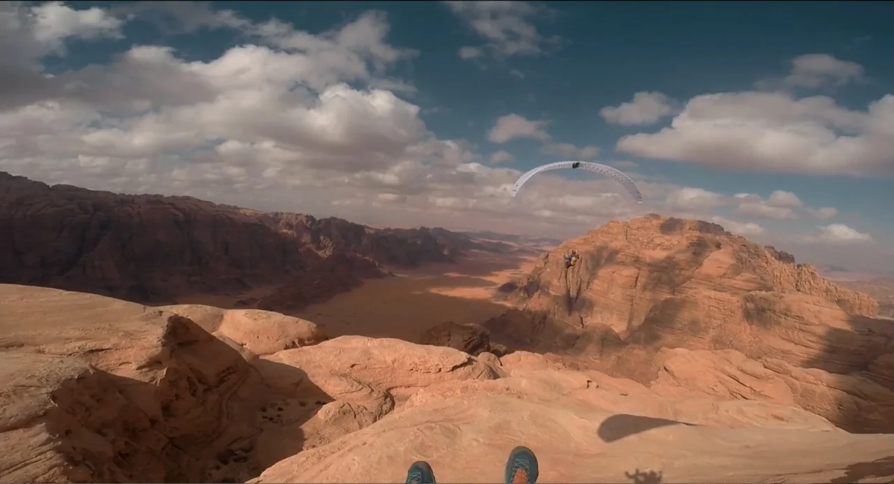 Crooked Trees Gon Give Me Wings (2022) movie still: wide — A vast rocky desert canyon spans out under a bright blue sky. A paraglider flies in the…
