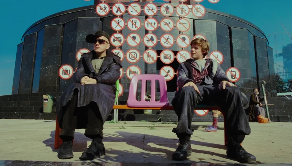 The Zero Theorem (2013) movie still: fullbody — A dark stone structure behind a bench displays many circular prohibition signs in red. A man and a…