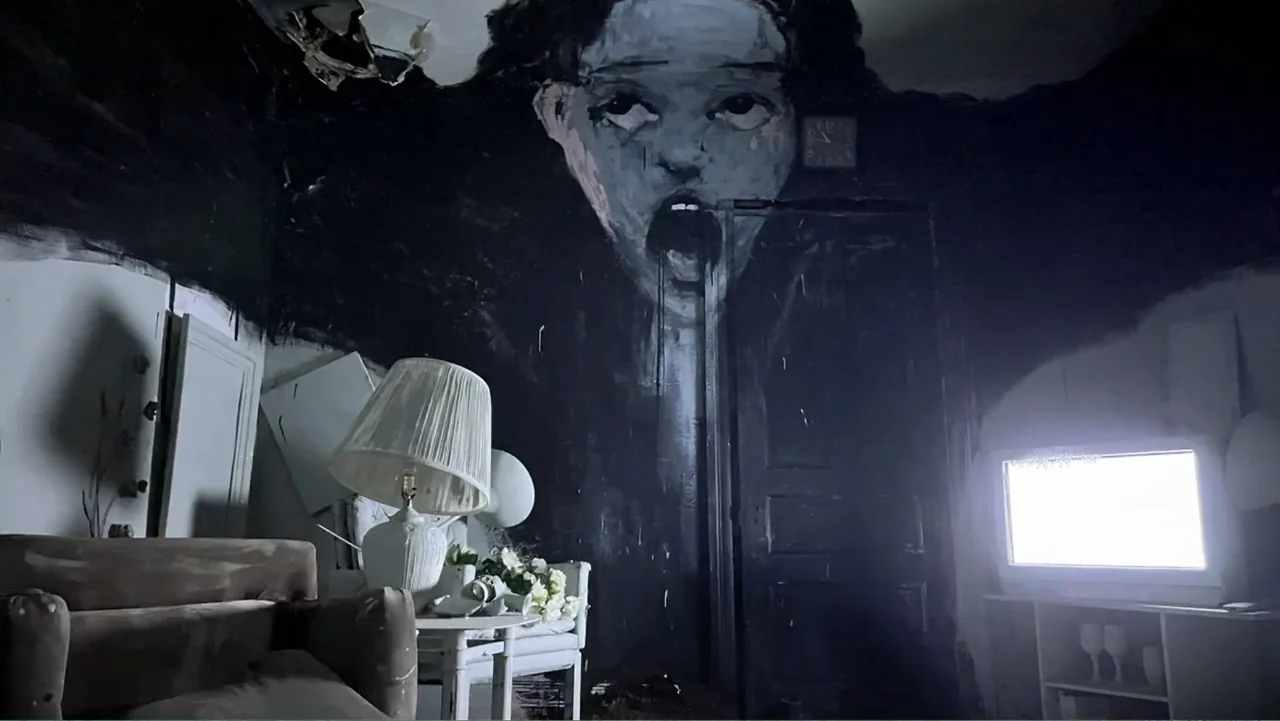 Inland Empire (2006) movie still: wide — A dimly lit room features a large black mural painted directly onto the wall. A bright television screen…