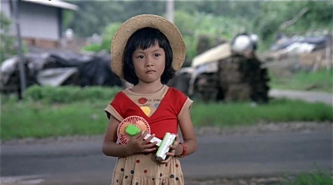 A Summer at Grandpa’s (1984) movie still: medium — A rural road paved with worn asphalt stretches past grassy verges and stacked wood. A young girl…