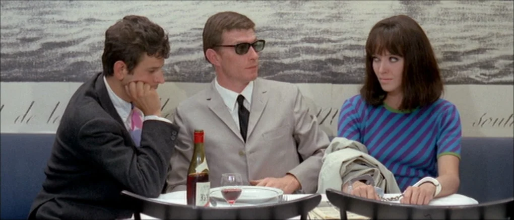 Made in U.S.A. (1966) movie still: medium — A French cafe interior features a table against a black-and-white landscape wall graphic. Three people…