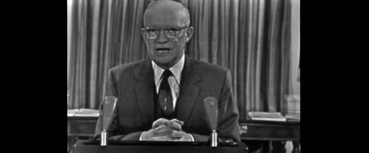 JFK (1991) movie still: medium — A formal office setting with vertically pleated curtains visible in the background. Dwight D. Eisenhower speaks…