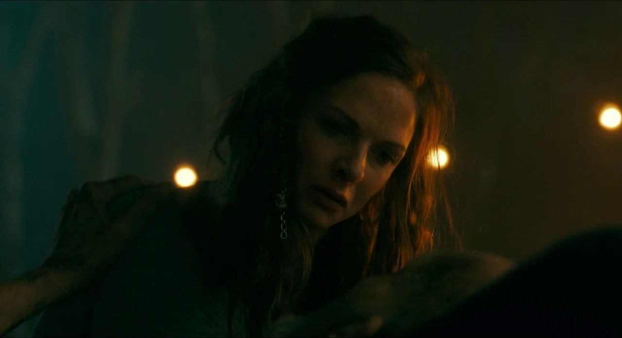 Doctor Sleep movie still: closeup — A dimly lit tent interior with a dark, blurred background containing several small light orbs. A woman with wavy…