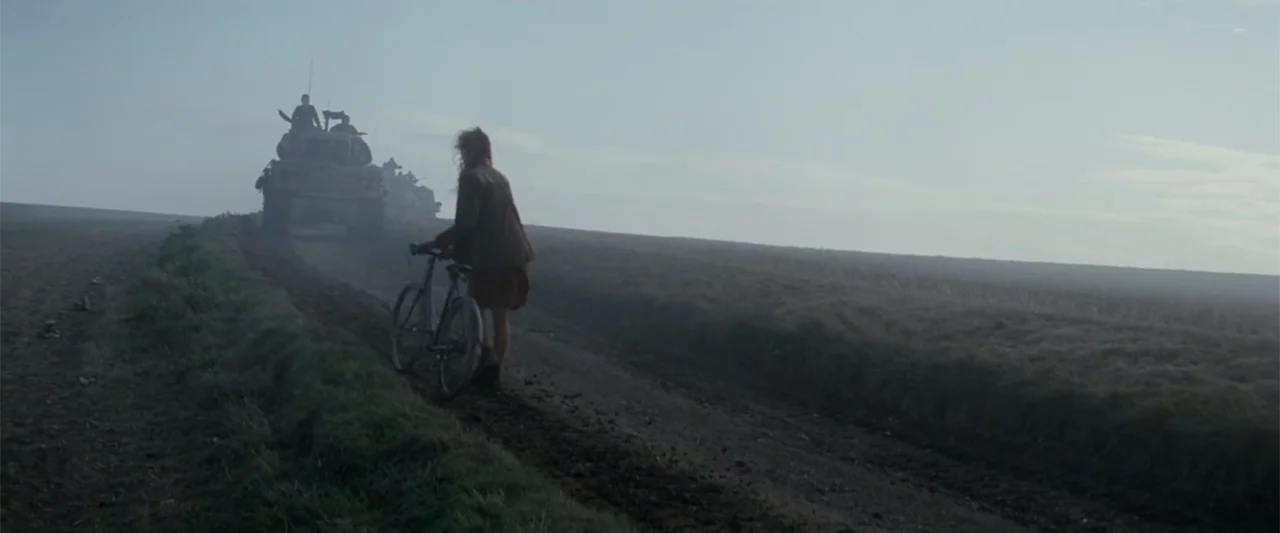 Fury (2014) movie still: wide — A wide, foggy dirt track cuts through an open, desolate field during the day. A woman stands in the middle of the…