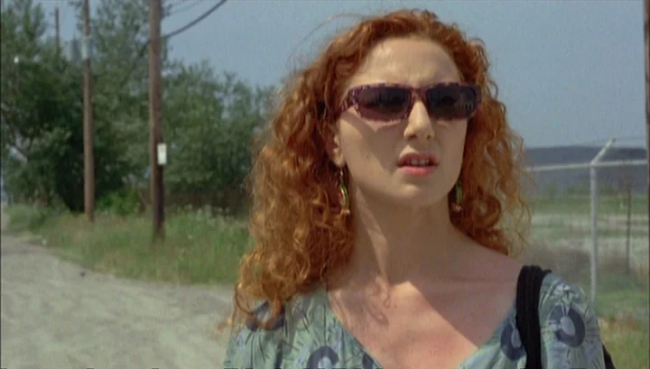 Highway 61 (1991) movie still: medium — A rural road with utility poles and a chain-link fence in the background. A woman with long curly red hair…