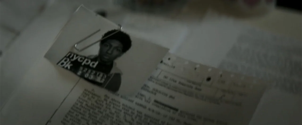 Crown Heights (2017) movie still: closeup — A close-up view shows a police mugshot photograph clipped to a stack of papers. The mugshot displays an…
