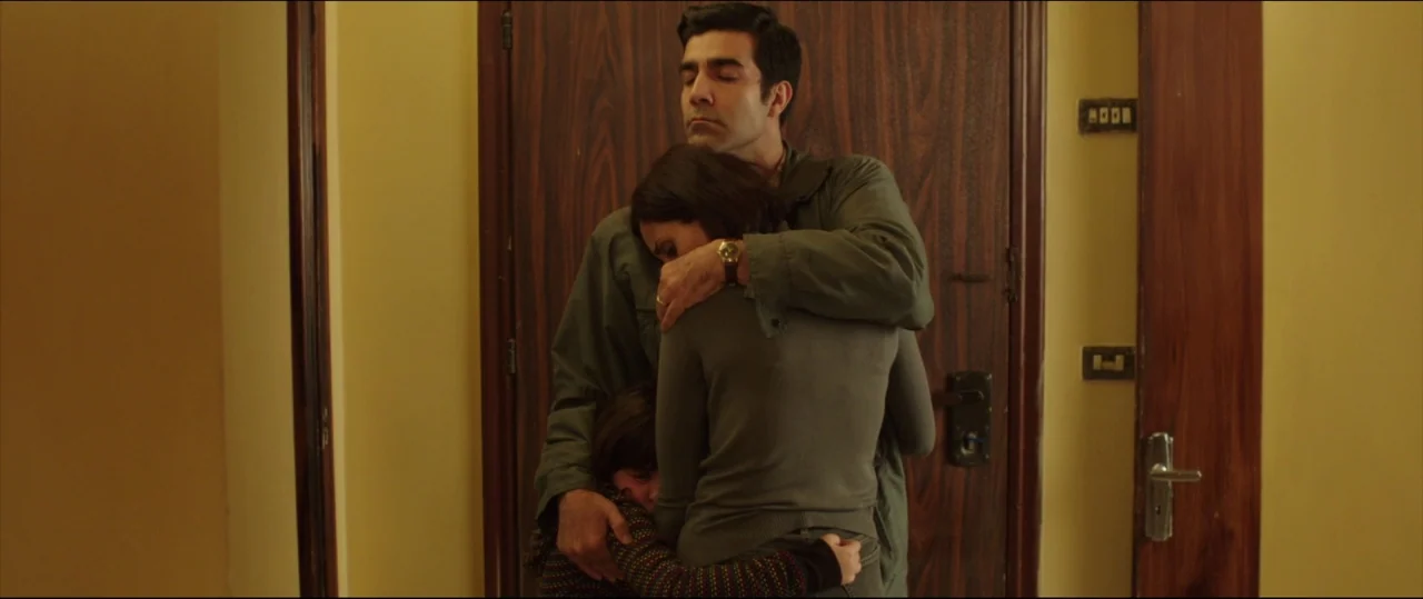Under The Shadow (2016) movie still: medium — A man and woman hug a small child in a narrow domestic hallway. The background features a wooden door…