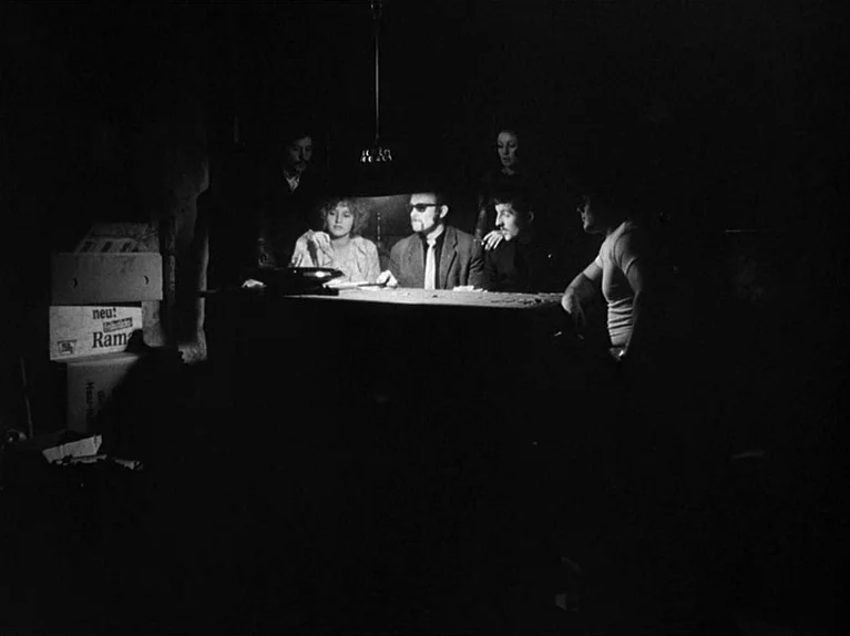 Gods Of The Plague (1970) movie still: medium — A dark, cluttered room with cardboard boxes stacked against a wall in the background. A group of…