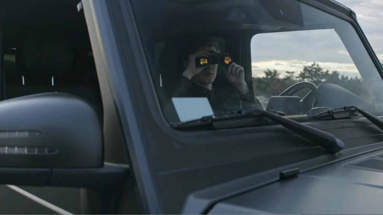 The Collective (2023) movie still: medium — A dark SUV interior viewed through the front windshield during the day. A young man sits in the driver…